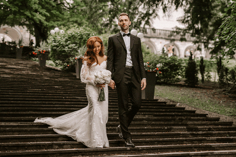 A beautiful bride from South Korea in a white fitted wedding dress with a simple white bouquet and the groom are holding hands and walking down the stairs in Villa Bled.