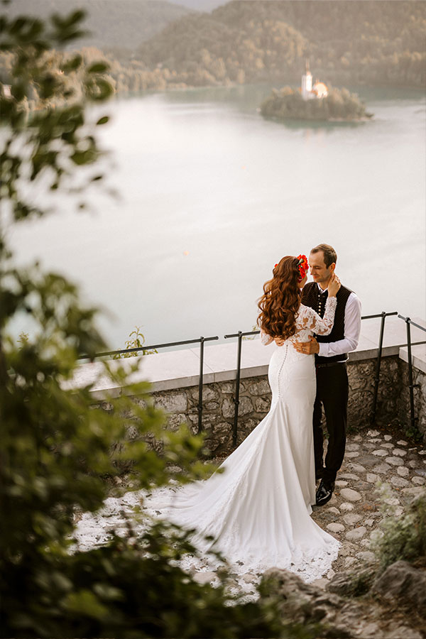 A close up photo of a bride and groom holding hands with a beautiful view of neighbury mountains, and a Bled lake with a famous island with Bled Church in the warm summer evening.