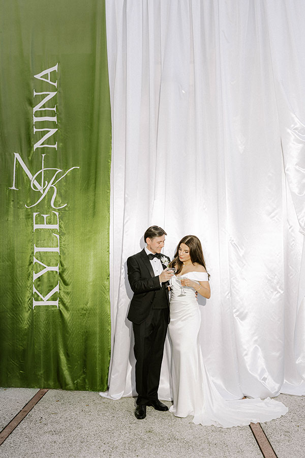 Bride and groom holding drinks in front of white draping and a green fabric panel personalised with their names.