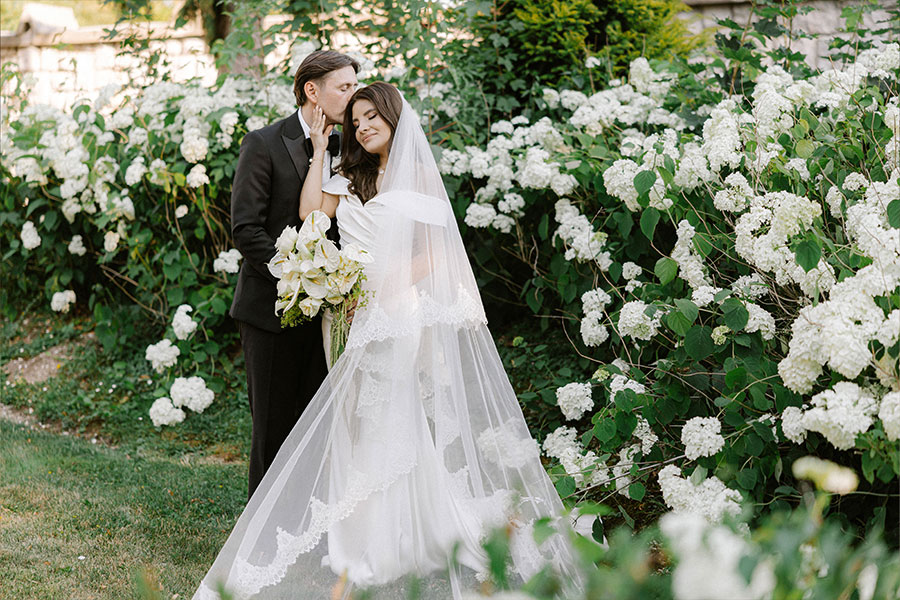 Groom kissing the bride’s forehead beside white hydrangeas, with the bride holding a white flower bouquet.