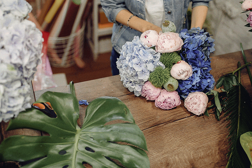 Wedding planner Petra Starbek in a flower shop making a bridal bouquet with greenery, pink peonies and blue hydrangea.