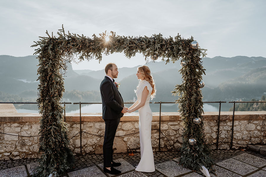 The bride in a stunning elegant dress and the groom in a black suit stand facing each other under a flower arch with Christmas lights at Lake Bled Castle.